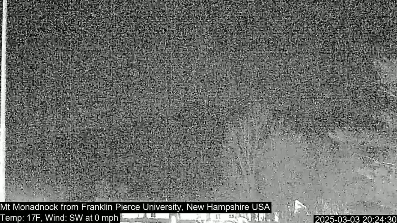 A nighttime, low-resolution image shows a dark, possibly snowy, landscape with indistinct shapes of trees and buildings under a cloudy sky.