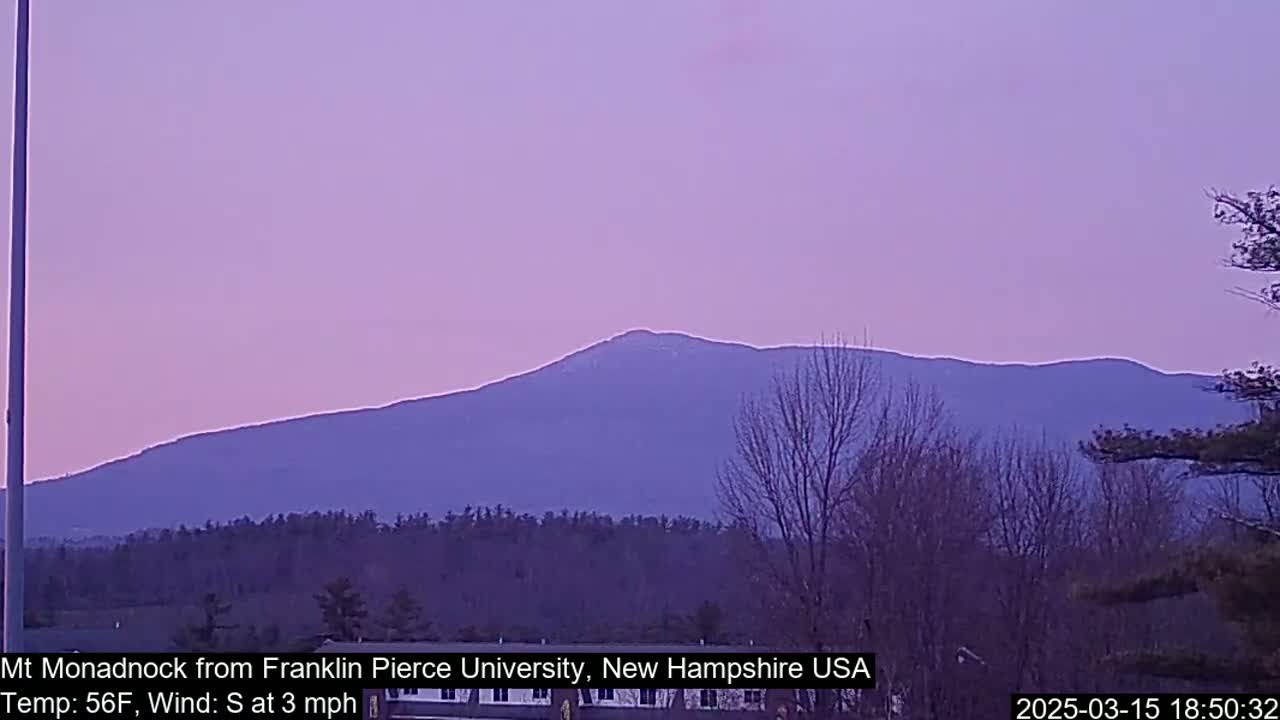 A distant, snow-capped mountain range is visible under a pale purple twilight sky, with bare trees in the foreground and a slight south wind.