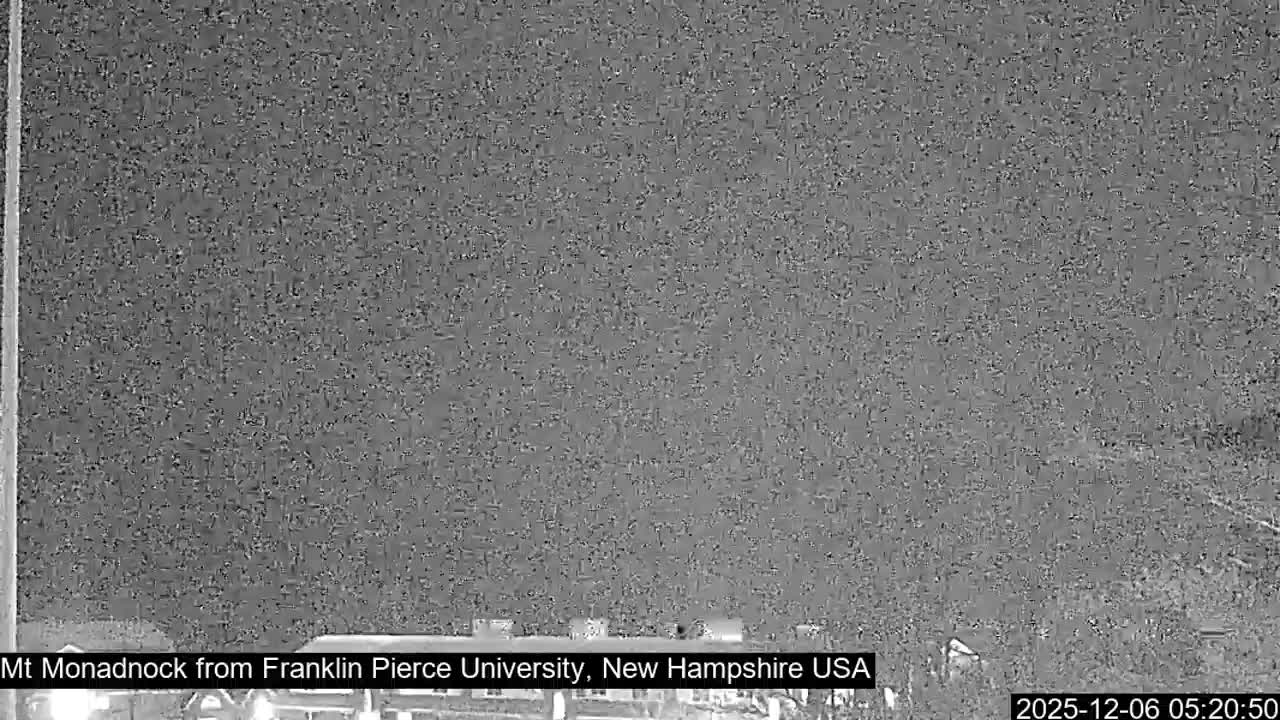 A heavily pixelated and noisy monochrome image captures a very dark outdoor scene at night, featuring faint, indistinct shapes at the bottom and a vertical element on the left, indicating extremely low visibility.