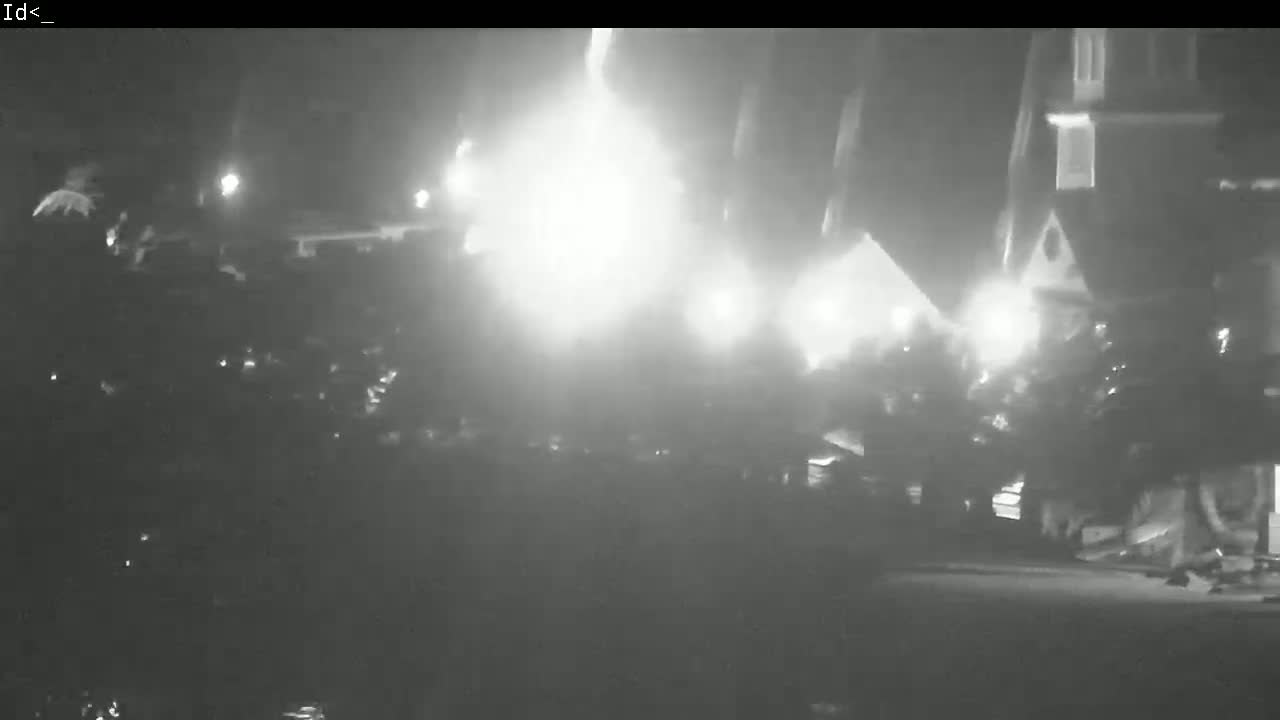 A nighttime, low-light image shows a bright, hazy fire engulfing a building and several people gathered nearby.