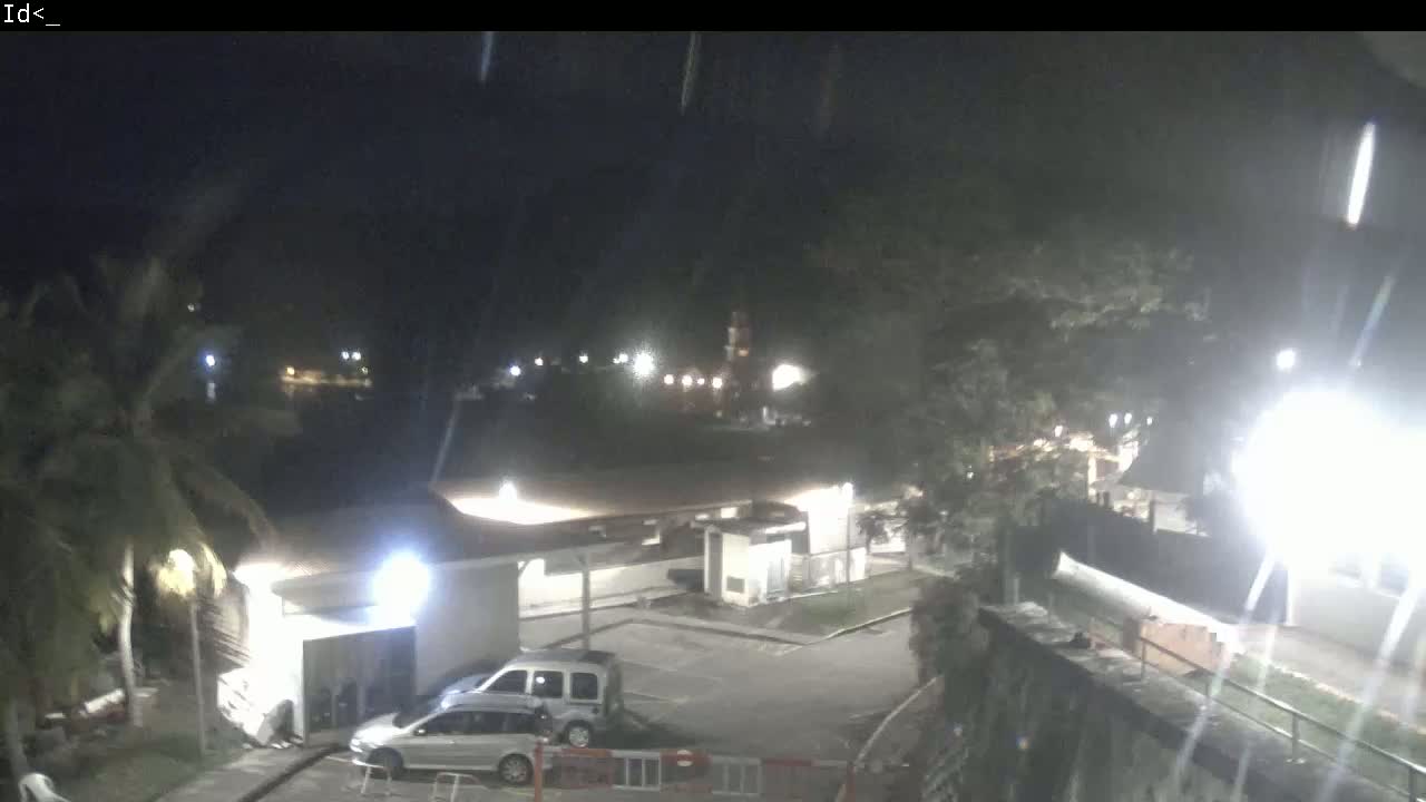 At night, under a dark sky with light rain, two vehicles are parked in a lot near a building surrounded by trees and lights.