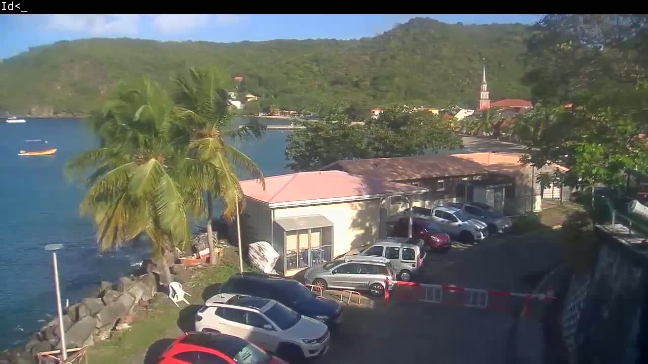 A waterfront scene on a sunny day shows several parked cars near a light-pink building, palm trees, and a body of water with a small boat, all set against a backdrop of lush green hills and a church steeple.