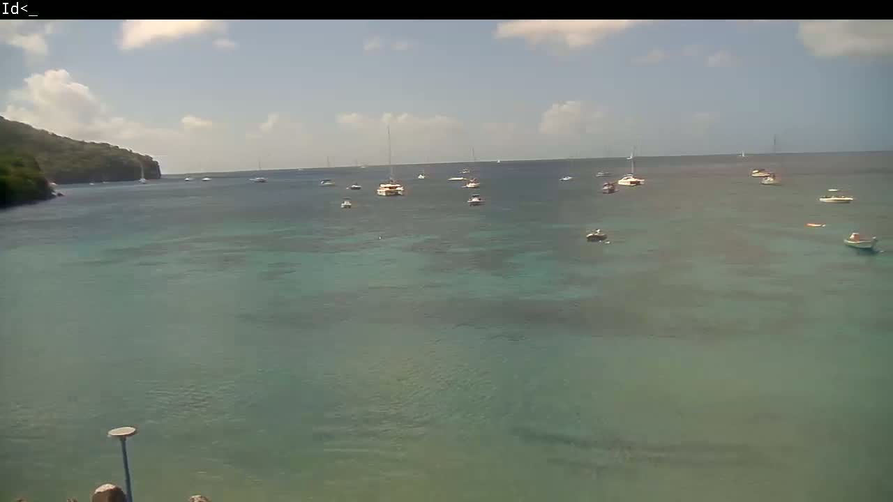 Several boats are anchored in a calm, teal-colored bay on a partly cloudy day, with a verdant hillside visible in the background.
