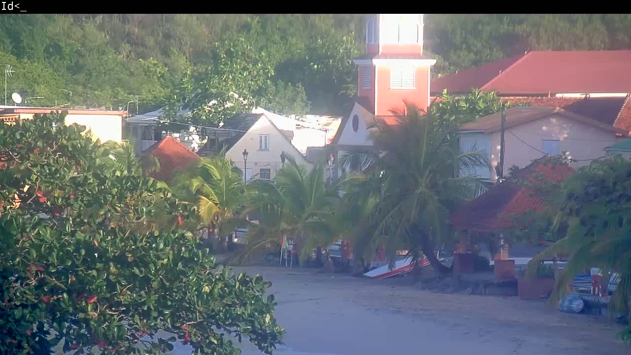 A sandy beach with tropical vegetation, buildings, and a church tower is seen on a sunny day.