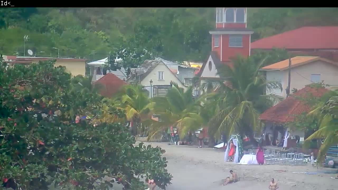 A beach wedding is taking place on a sandy shore in front of tropical buildings under an overcast sky.