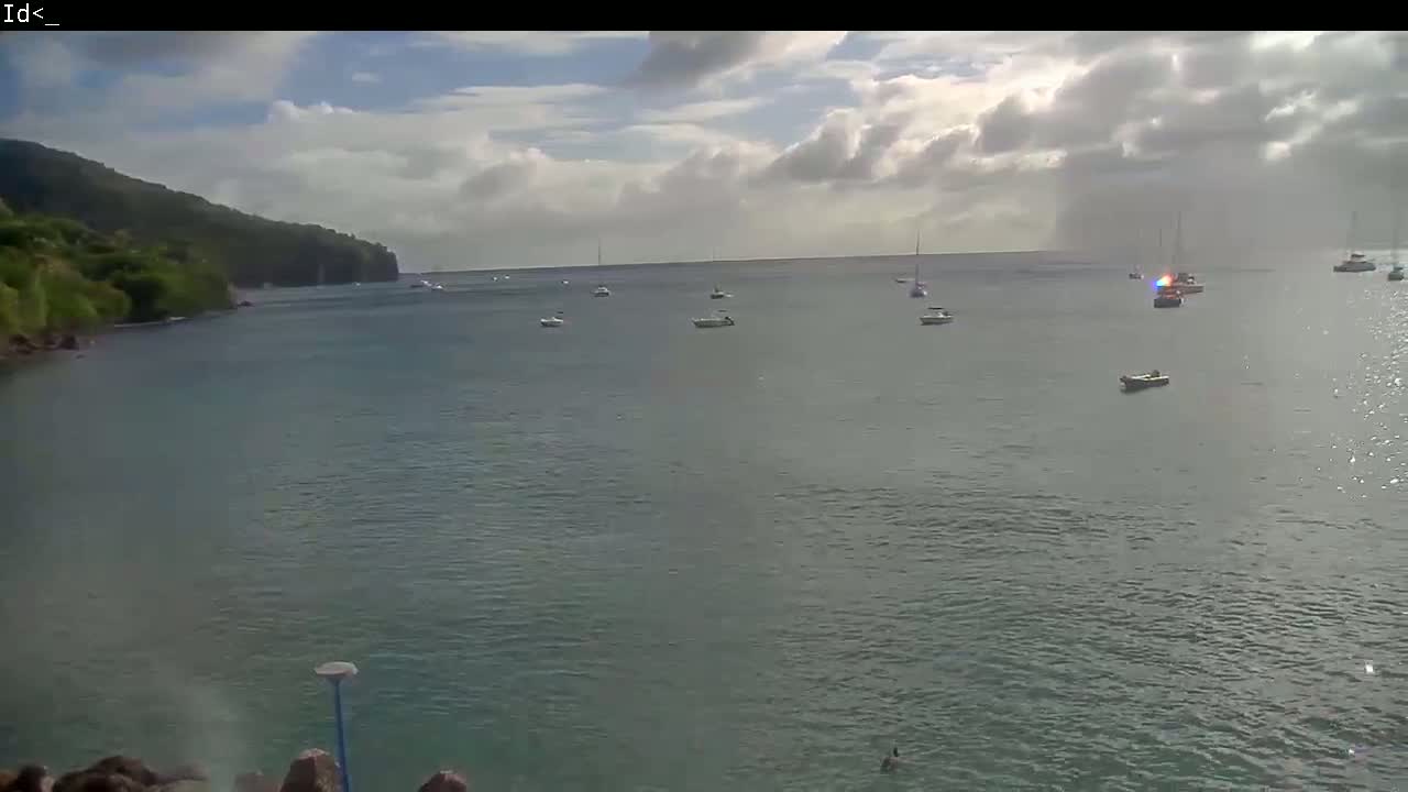 A calm bay with several boats is viewed from a rocky shore under a partly cloudy sky.
