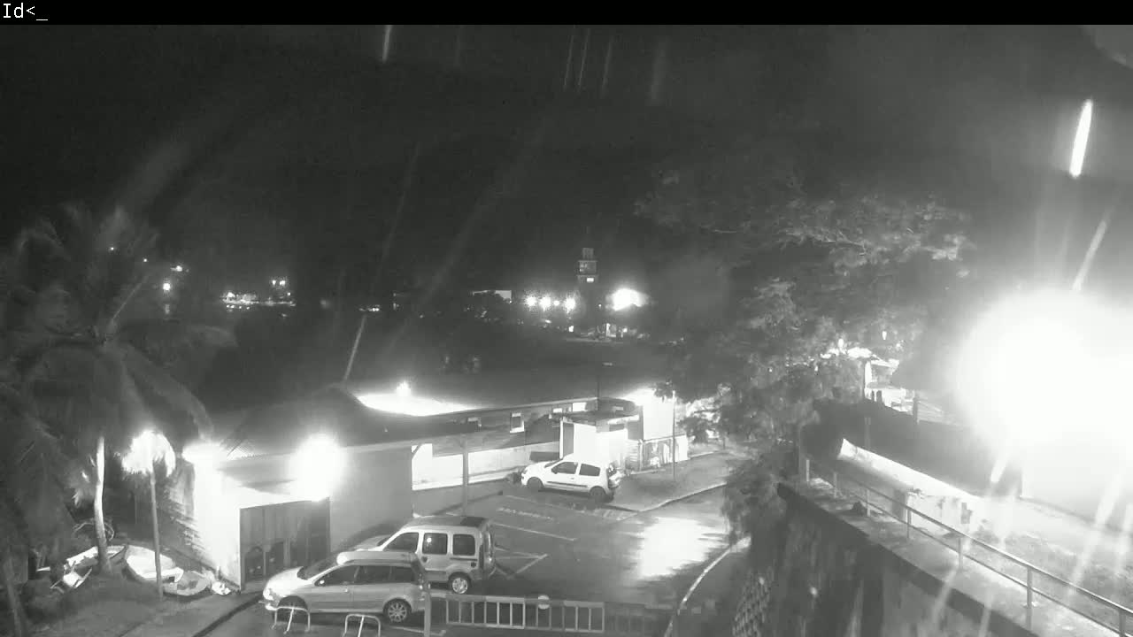 A nighttime, rain-swept view shows a parking lot with several cars near a low building, all under dimly lit conditions.