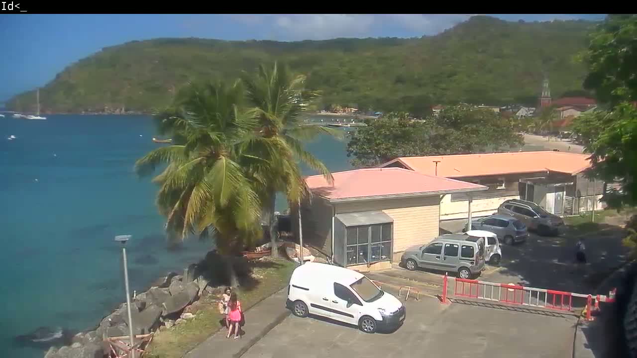 A sunny, tropical scene shows a calm bay with sailboats, palm trees, a row of low buildings, and parked cars near a rocky shoreline.