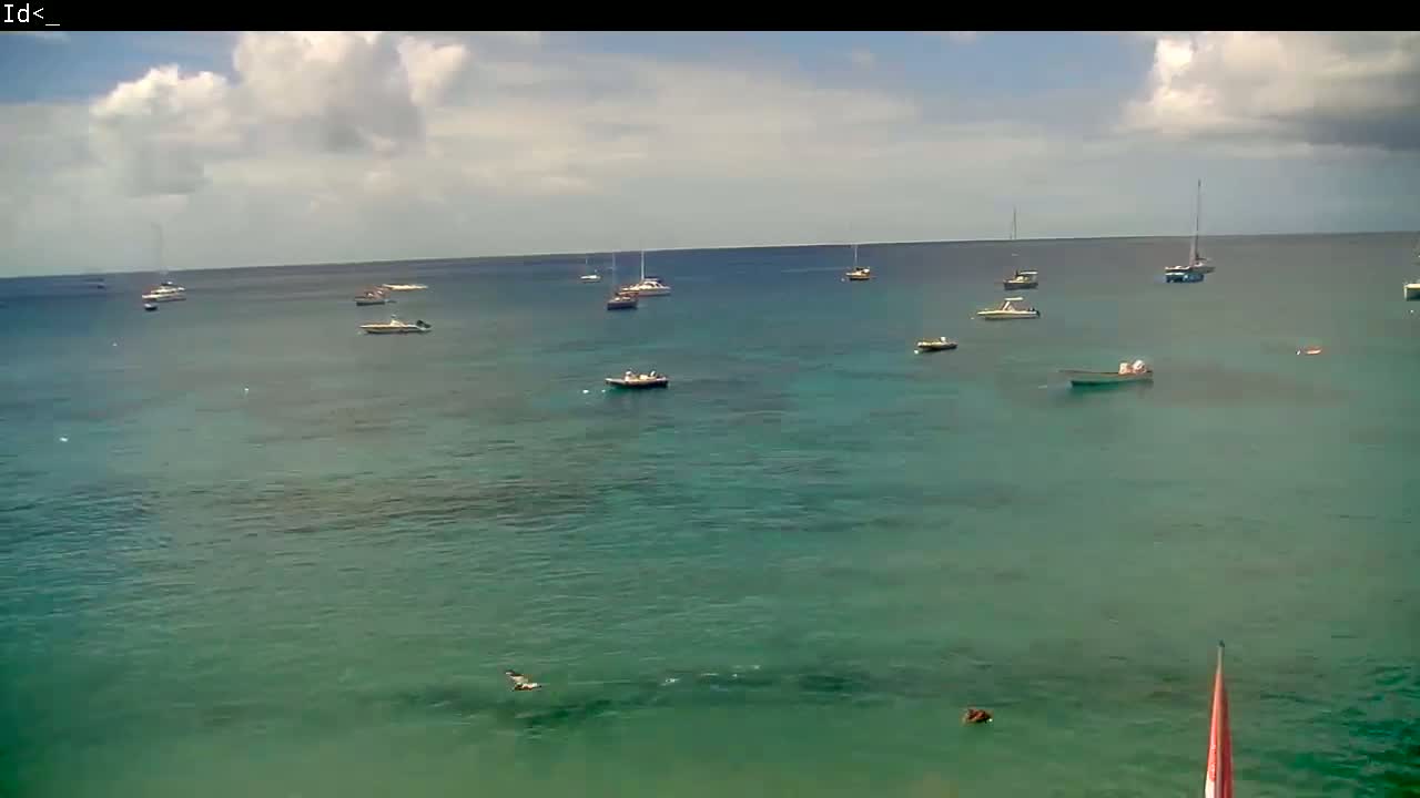 A partly cloudy day shows several boats and a few people swimming in calm, teal-colored water.