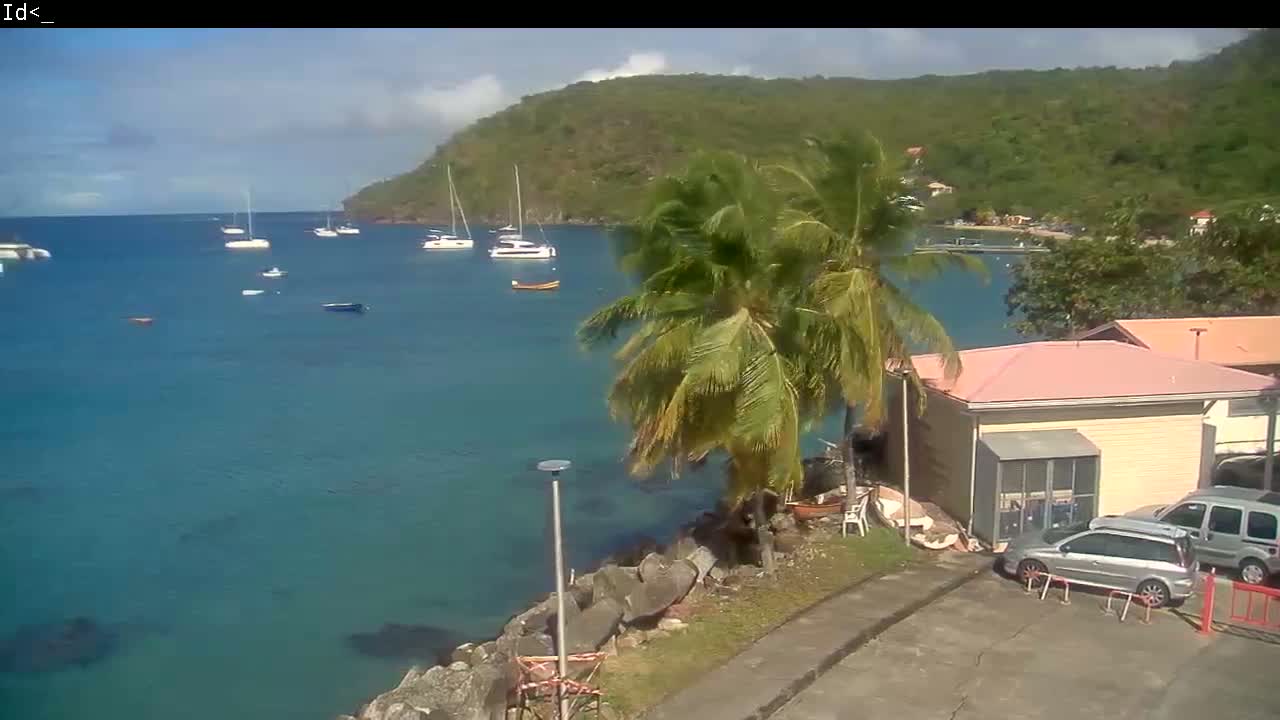 A calm, teal bay with several sailboats anchored, is viewed from a high vantage point near a palm tree-lined shore with a small building and parked cars, under a partly cloudy sky.