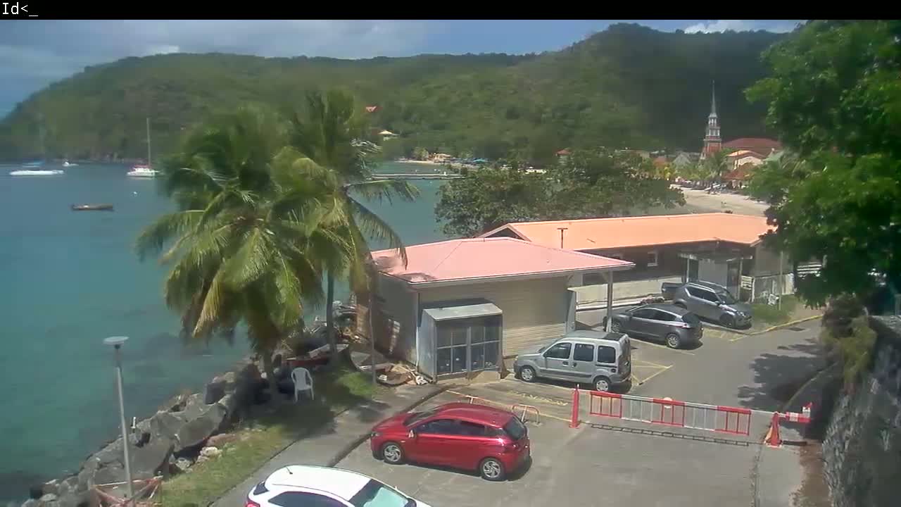 A partly sunny day reveals a seaside town nestled beside a calm bay, featuring a pink building, several parked cars, and lush tropical vegetation including palm trees.