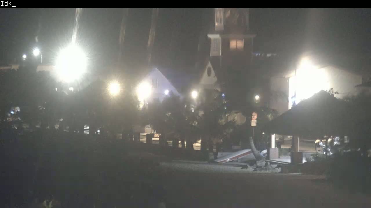 At night, brightly lit buildings and a thatch-roofed structure are visible, with some people gathered near the structure under hazy conditions.