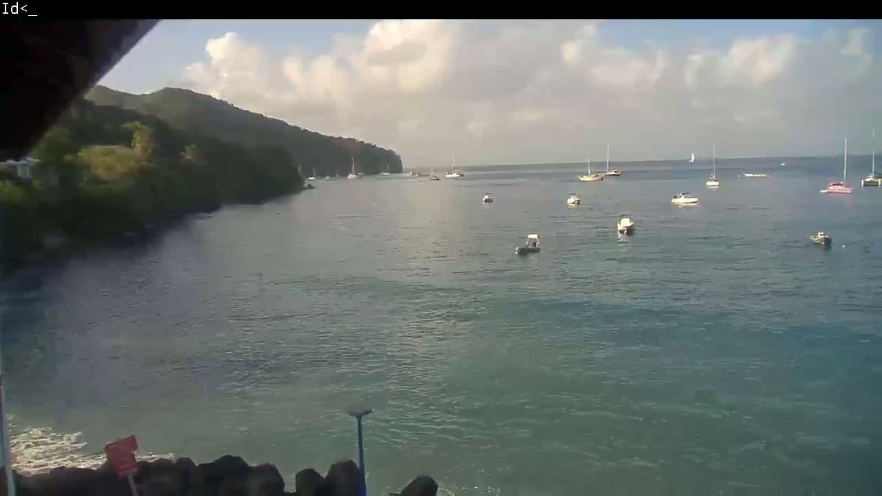 A calm, partly cloudy day shows a bay with numerous boats and sailboats anchored near a lush, green hillside.