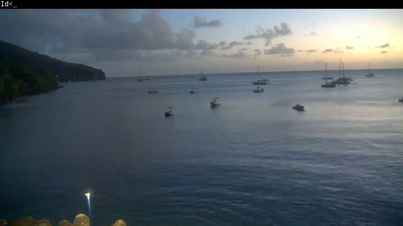 Several boats and sailboats are anchored in a calm bay at dusk, with a dark, forested shoreline visible in the background.