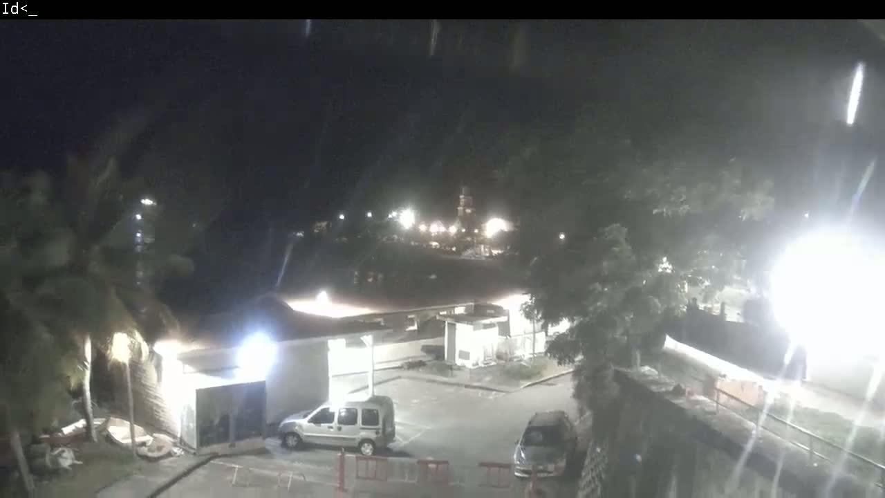 A nighttime scene of a parking lot with two vehicles, surrounded by buildings and trees under a light rain.