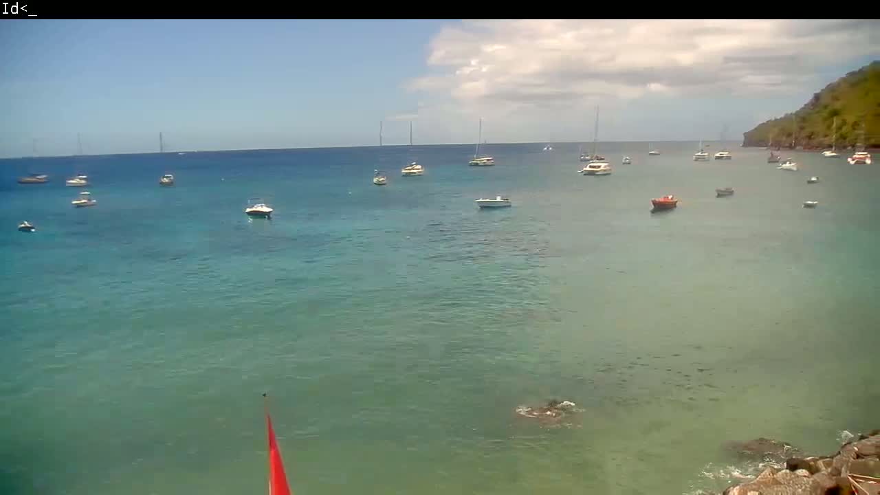 A partly cloudy day shows numerous boats anchored in a calm, clear teal ocean near a verdant hillside.