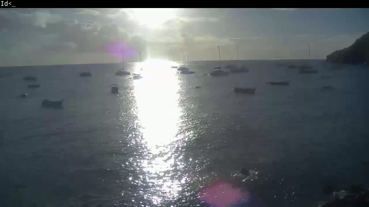 Several boats are moored in calm water under a partly cloudy sky, with the sun shining brightly on the water's surface.