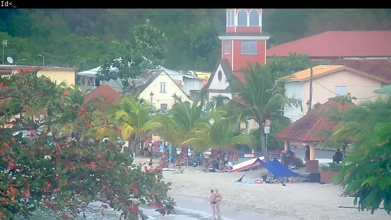 A beach scene shows a sandy shoreline with people relaxing, several buildings and palm trees behind, all under an overcast sky.