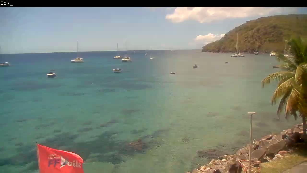 A partly cloudy day shows a calm, clear turquoise bay with numerous sailboats and motorboats anchored, a rocky shoreline, and a lush green hill in the background.