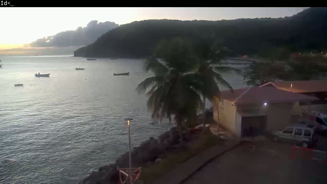 Several small boats are in calm water near a palm tree and a building at sunset under a partly cloudy sky, with hills in the background.