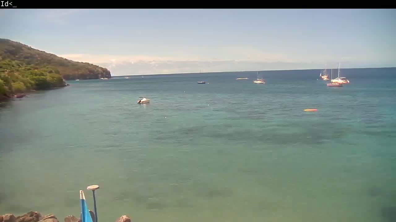 A calm, clear ocean bay is seen from a rocky shoreline, with several boats anchored in the distance and a green hillside visible to the left.