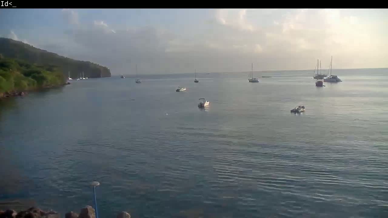 Several boats are anchored in a calm bay on a partly cloudy day, with a green hill visible in the background.
