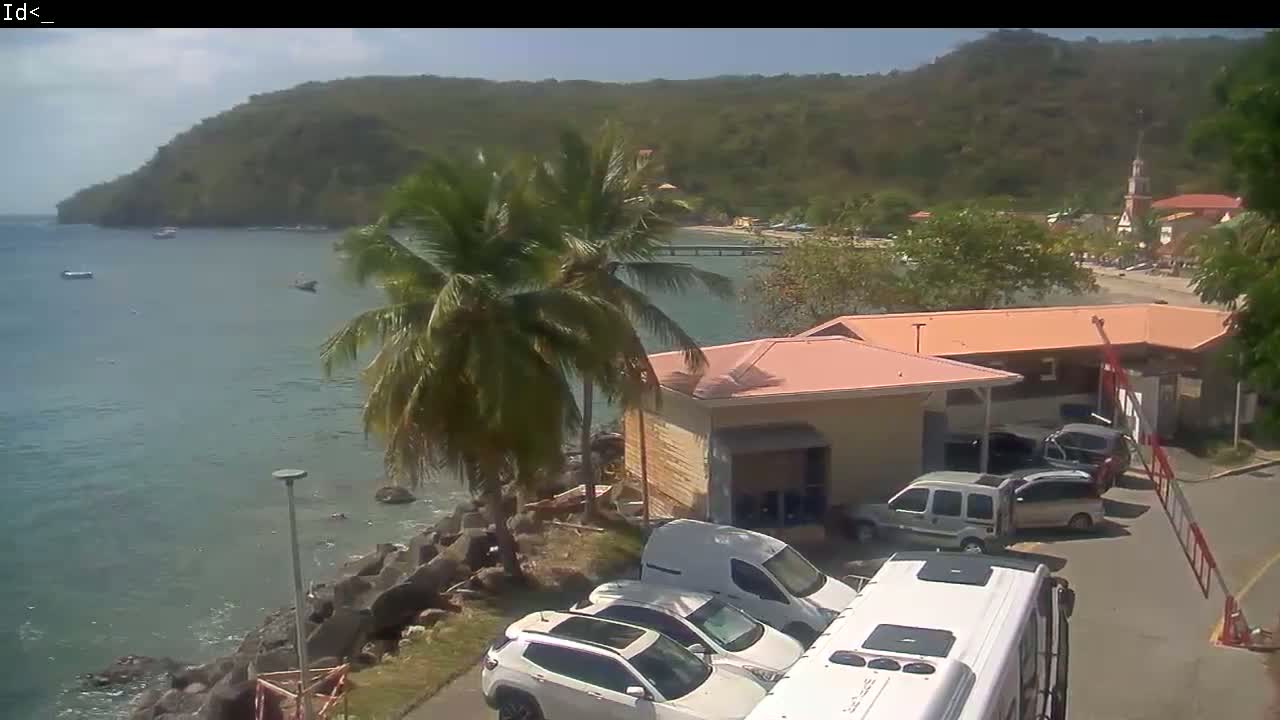 A tranquil waterfront scene on a sunny day shows a parking lot with several cars and a van near buildings, with a calm body of water and green hills in the background.