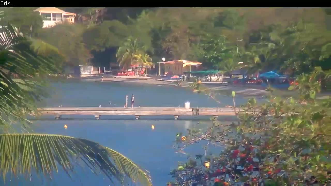 A calm body of water with a pier where two people are walking, is surrounded by lush green vegetation, and several buildings are visible in the background on a sunny day.