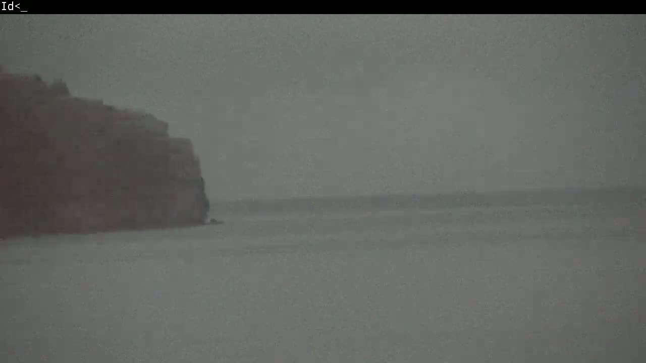 A dark, overcast day shows a rocky cliff overlooking a calm, gray body of water.