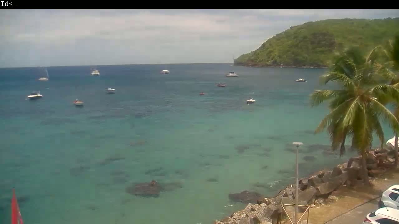A calm, clear turquoise bay with several boats at anchor, a green hillside in the background, and palm trees near a rocky shoreline on a partly cloudy day.