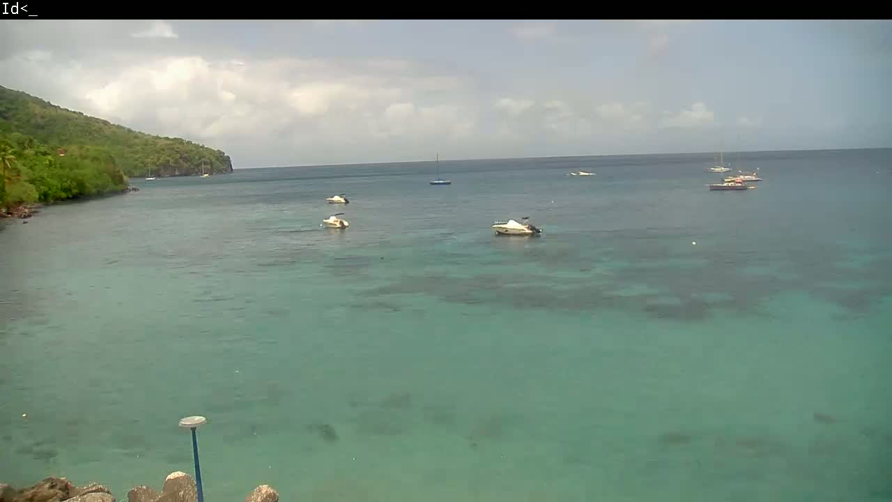 Several boats are anchored in a calm, teal-colored bay, partially sheltered by a verdant hillside under a partly cloudy sky.