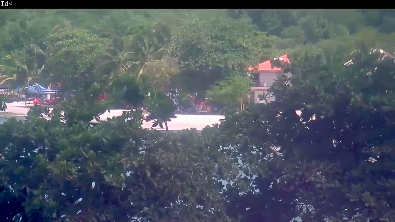 A light-colored building with a red roof is partially visible behind lush green vegetation, overlooking a sandy beach area with a few people and a small structure.
