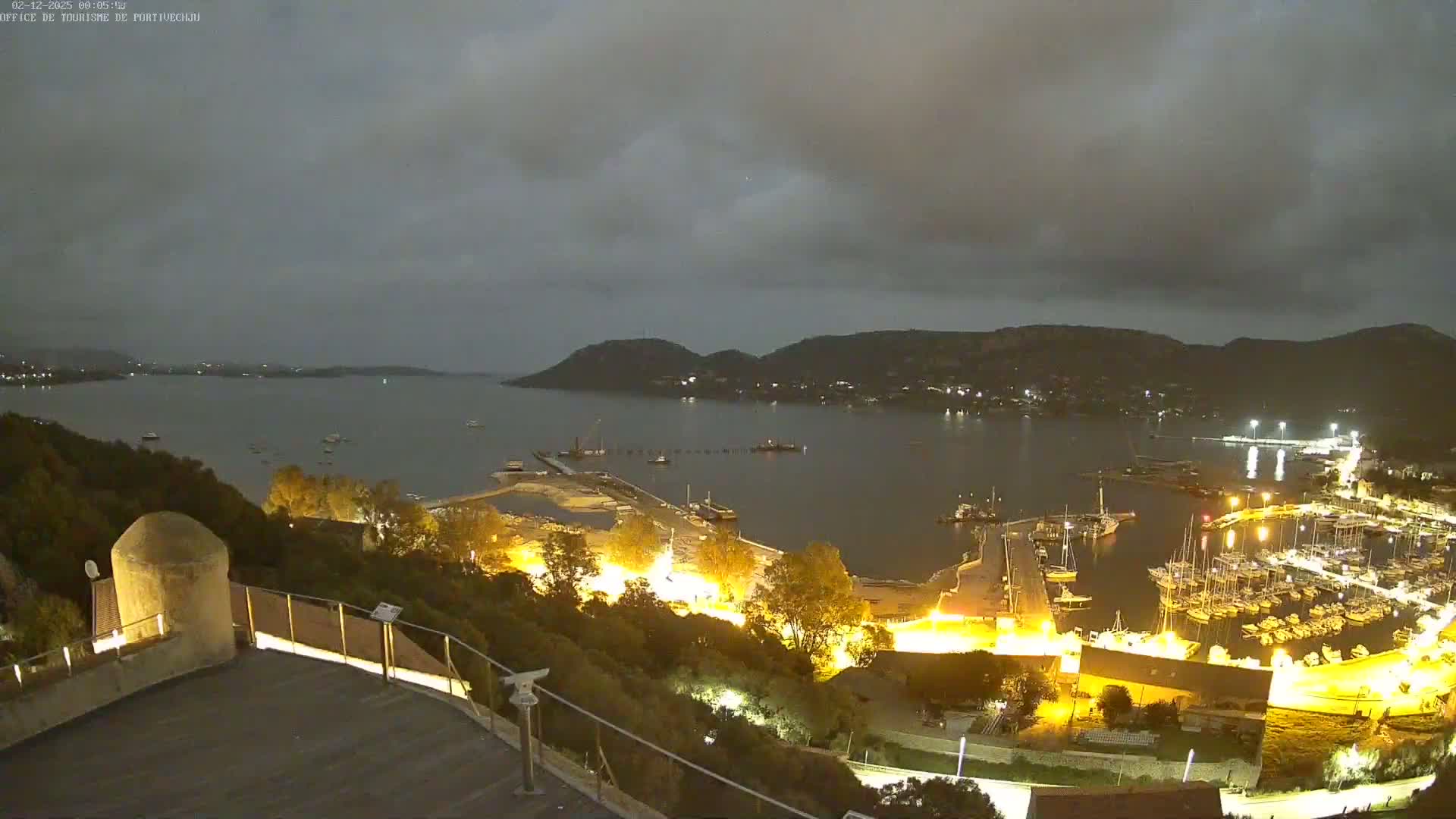 A nighttime view of a harbor filled with boats, illuminated against a backdrop of dark, cloudy skies and hills.