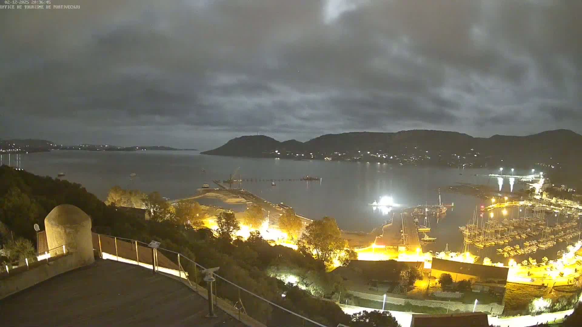 A nighttime view of a harbor filled with boats, under a cloudy sky, shows a coastline with hills and lights illuminating the area.