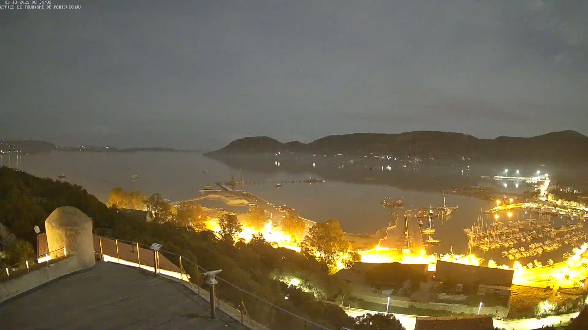 A nighttime view overlooking a calm harbor filled with boats, surrounded by hills and lit up with many lights.