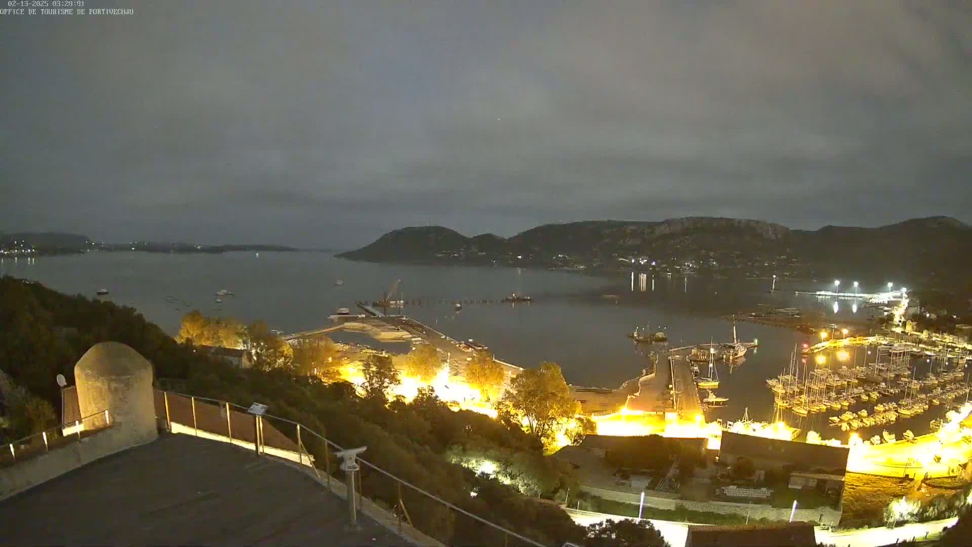 A nighttime view of a harbor filled with boats, illuminated buildings, and a calm bay under a cloudy sky.