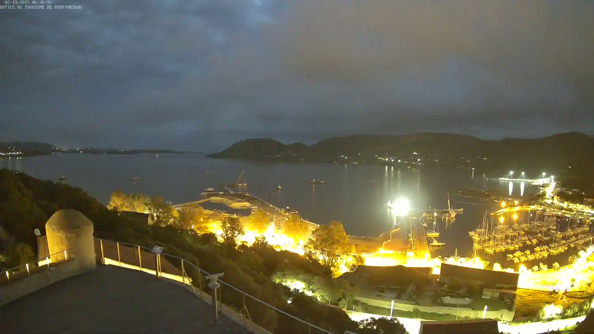A nighttime view from an elevated vantage point shows a harbor filled with boats, lit up against a dark, cloudy sky, with hills visible in the background.