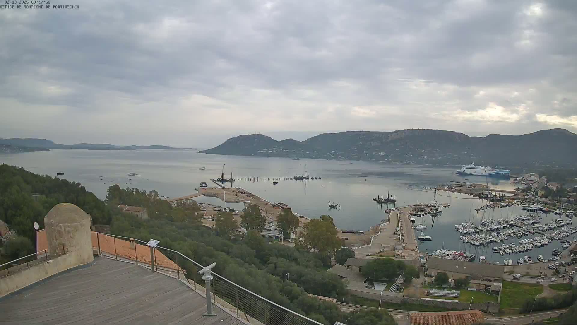 A high-angle, long shot of a harbor filled with boats under an overcast sky, with construction visible near the water and hills in the background.