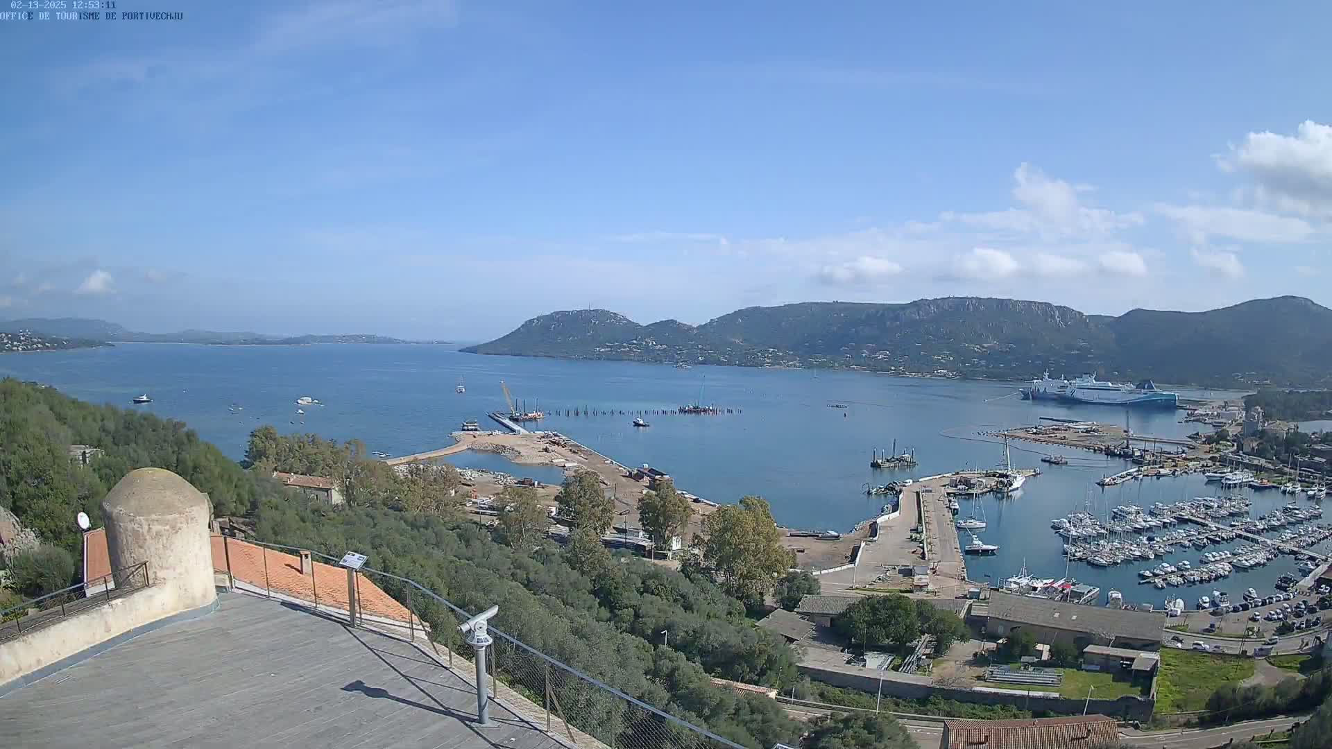 An elevated view of a harbor filled with numerous boats and ships, under a mostly clear sky, with mountains in the background.
