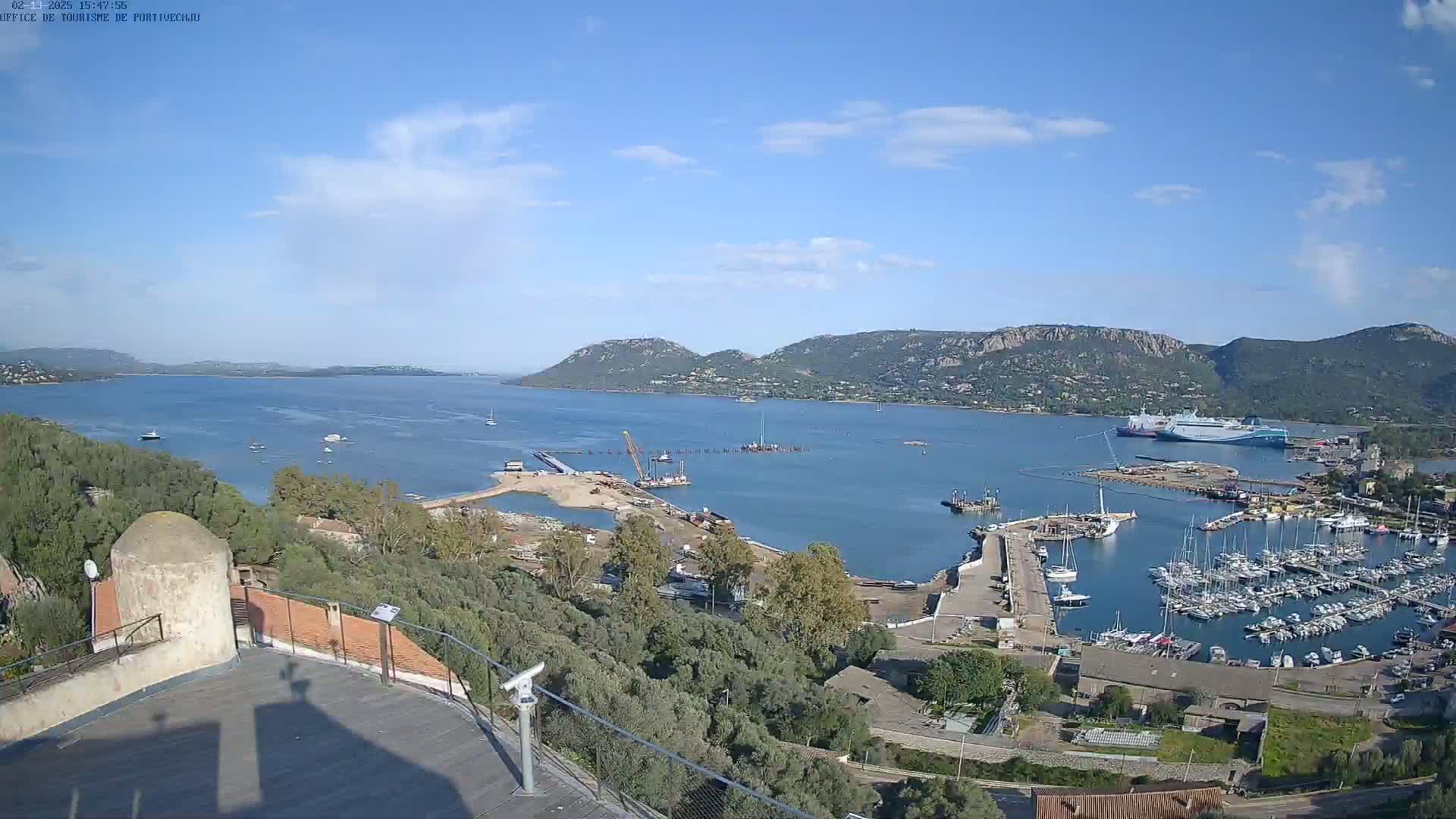A partly sunny day overlooking a harbor with many boats, construction activity, and a large ferry docked, all situated against a backdrop of hills and a calm bay.