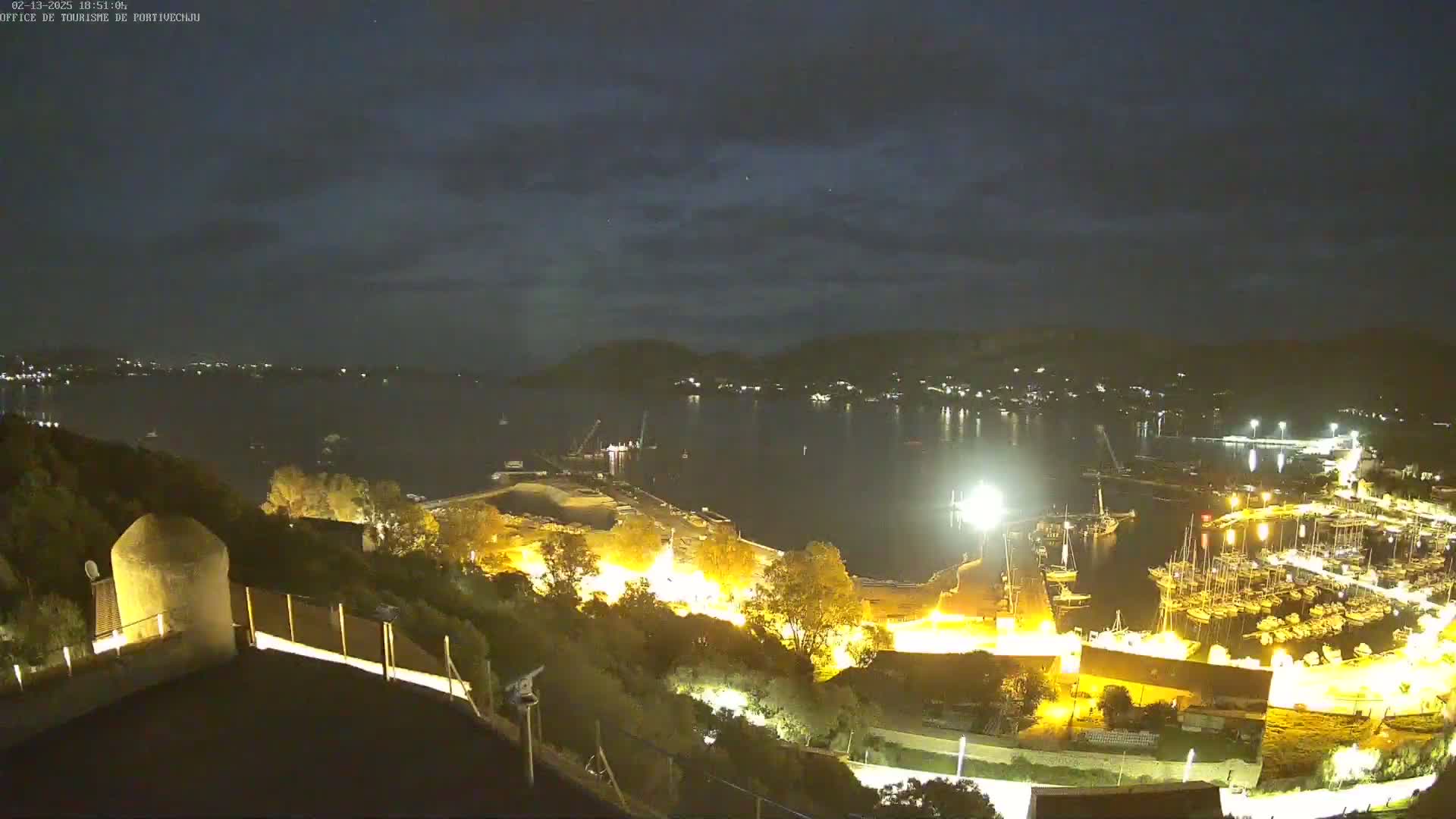 A nighttime view overlooking a harbor filled with boats, illuminated by artificial lights under a partly cloudy sky.