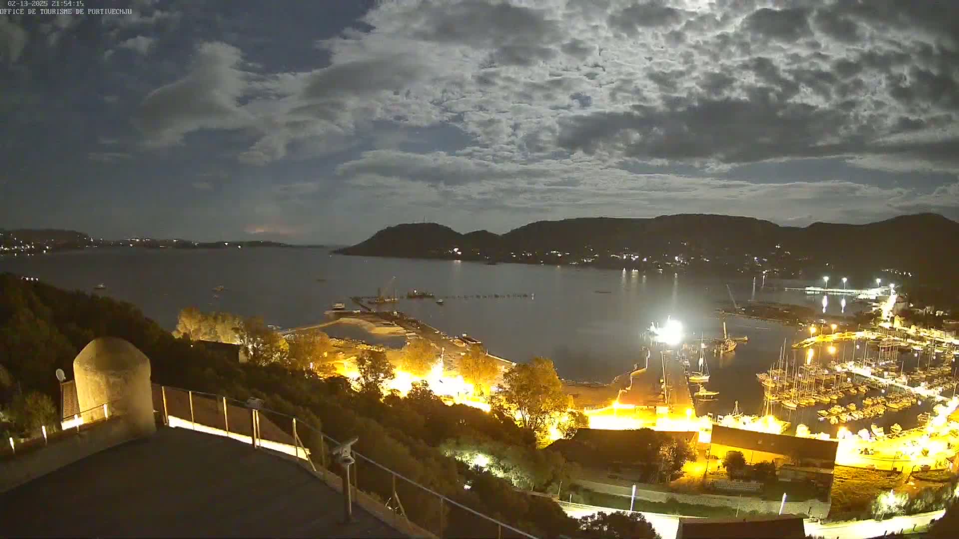 A nighttime view of a harbor filled with boats, illuminated by lights, under a partly cloudy sky with a bright moon.