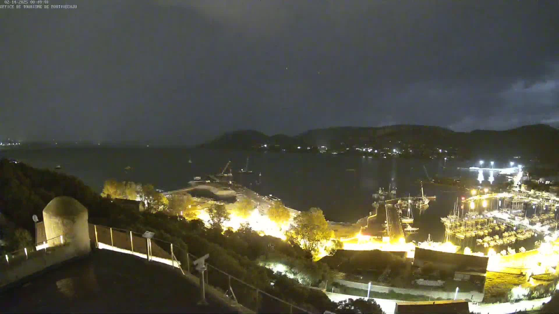 A nighttime, high-angle view shows a harbor filled with boats, illuminated buildings along the shore, and hills in the background under a dark, cloudy sky.