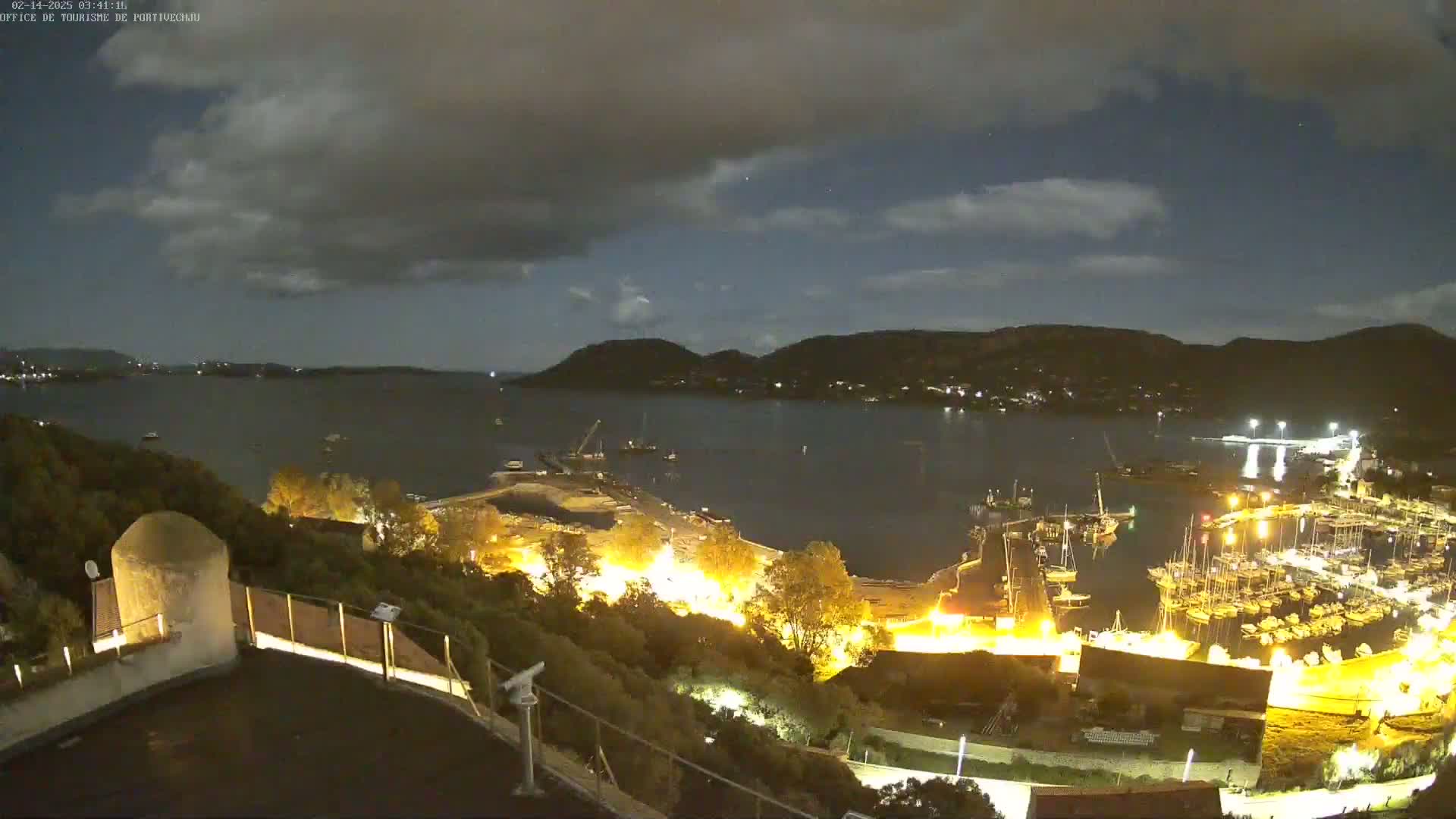 A nighttime view of a harbor filled with boats, illuminated against a backdrop of hills under a partly cloudy sky.