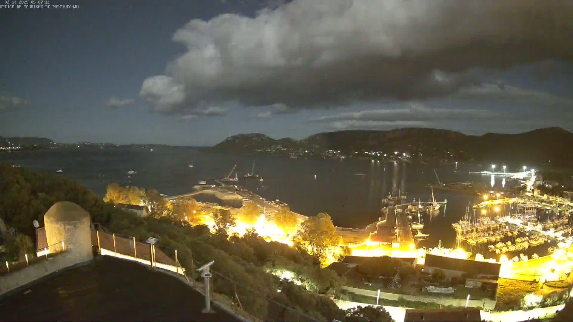 A nighttime view of a harbor filled with boats, illuminated buildings, and hills under a partly cloudy sky.
