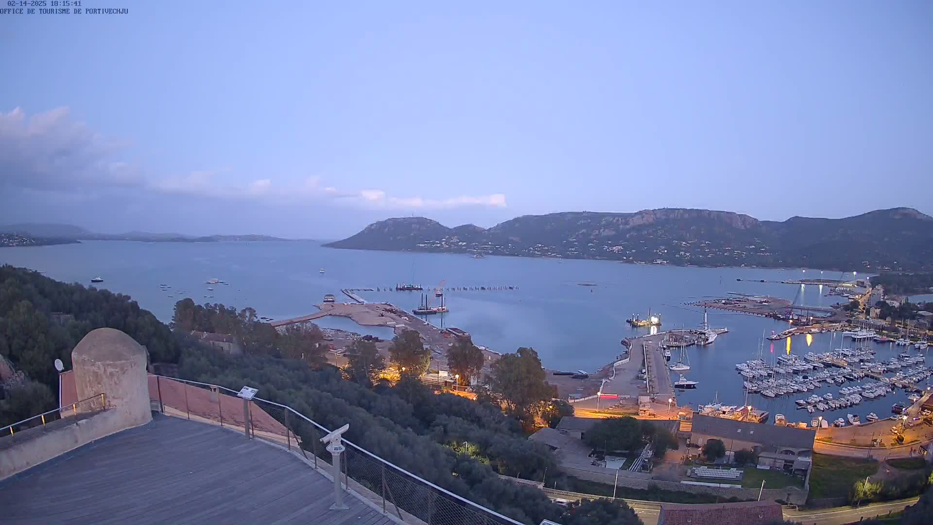 An elevated view at twilight shows a calm bay with a marina filled with boats, construction activity near the water, and hills in the background under a clear sky.