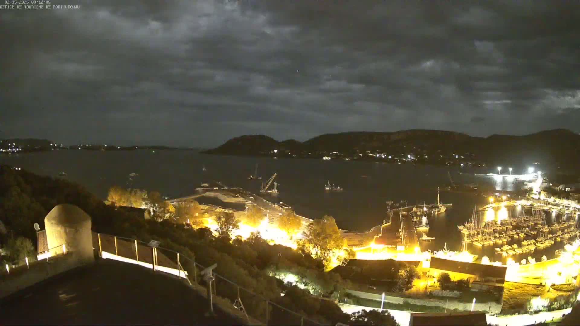 A nighttime view of a harbor filled with boats, under a cloudy sky, as seen from a hilltop vantage point.