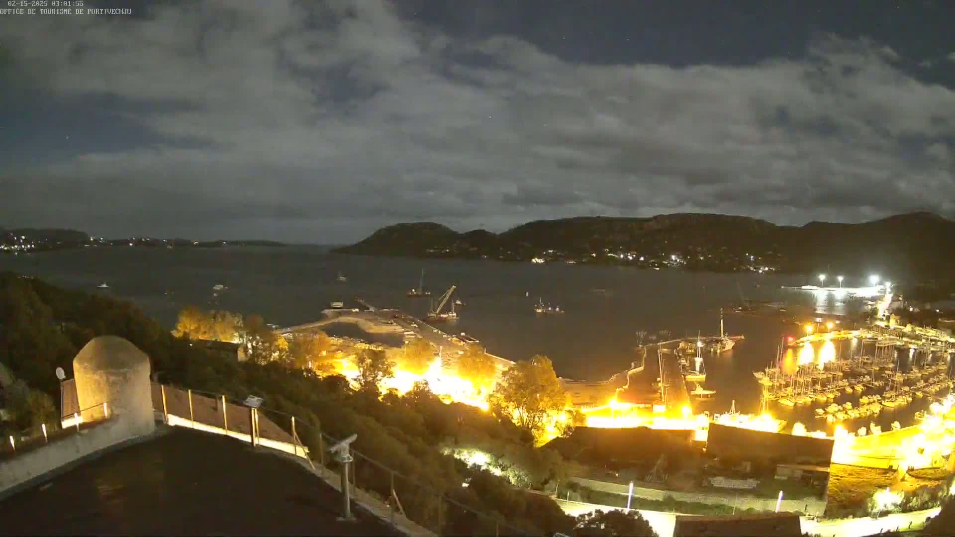 A nighttime view of a harbor filled with boats, illuminated by lights, under a partly cloudy sky.
