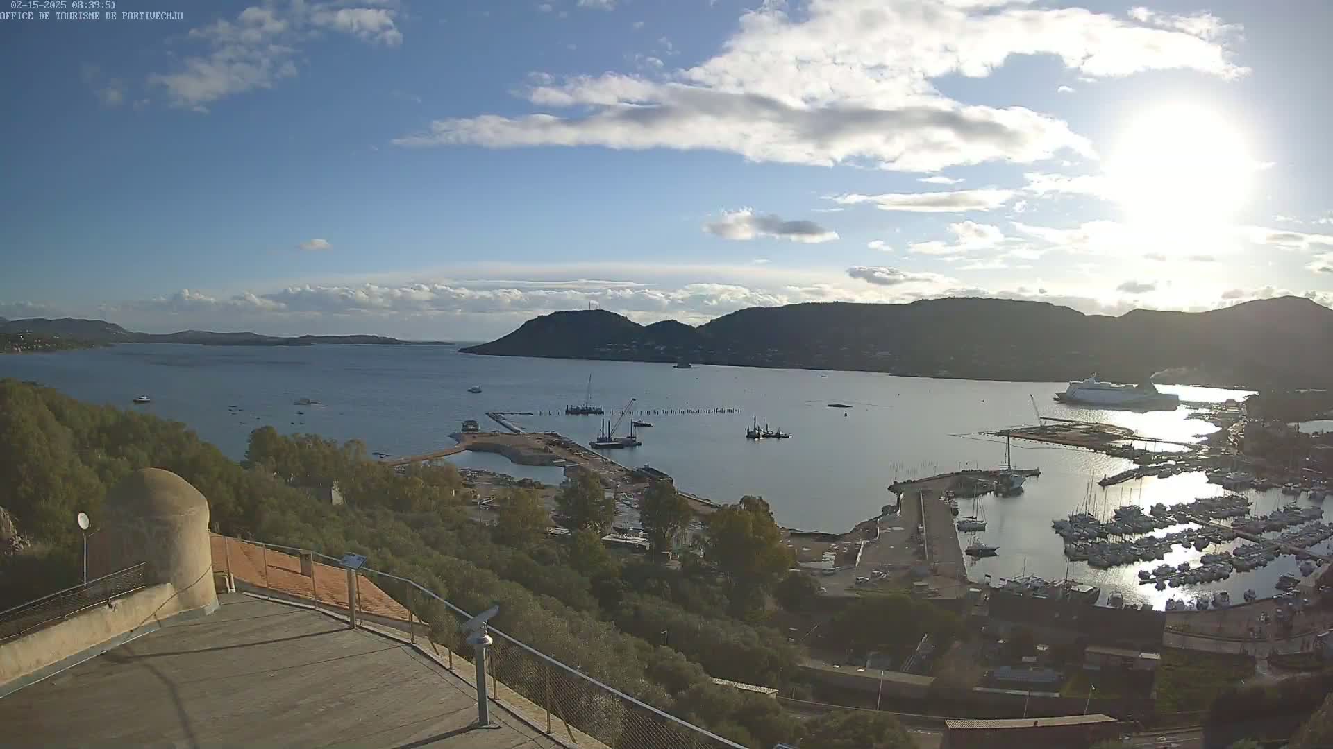 A partly sunny day overlooks a harbor filled with many boats and a large ferry, nestled between hills and a calm body of water.