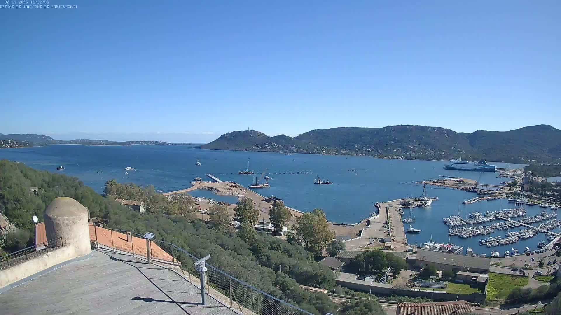 A clear, sunny day overlooks a harbor filled with boats and a large ferry, nestled between hills and a calm body of water.